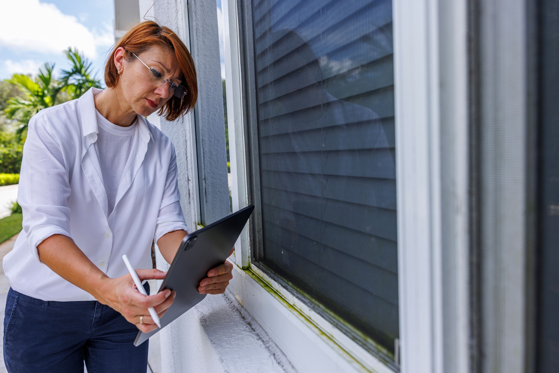 Windows leakage inspection with digital tab: red haired claim agent inspecting windows for signs of leakage, water stains or mold outdoors